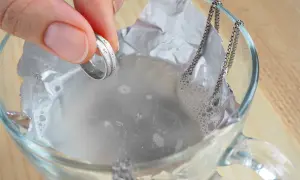 Dłoń trzyma pierścionek nad szklanką z wodą i folią aluminiową, obok wisi łańcuszek.