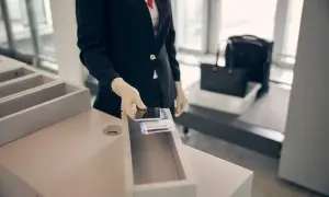 Pracownik lotniska w rękawiczkach wkłada telefon i bilety do pojemnika. Bagaż podręczny czeka na prześwietlenie.