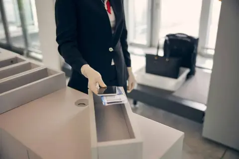 Pracownik lotniska w rękawiczkach wkłada telefon i bilety do pojemnika. Bagaż podręczny czeka na prześwietlenie.