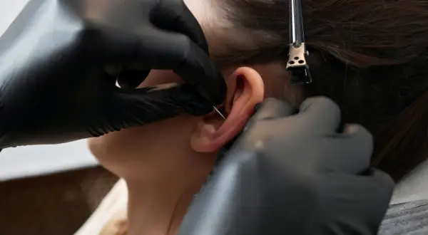 Profesjonalne wykonanie kolczyka orbital piercing. Ręce w czarnych rękawiczkach precyzyjnie przekłuwają ucho.