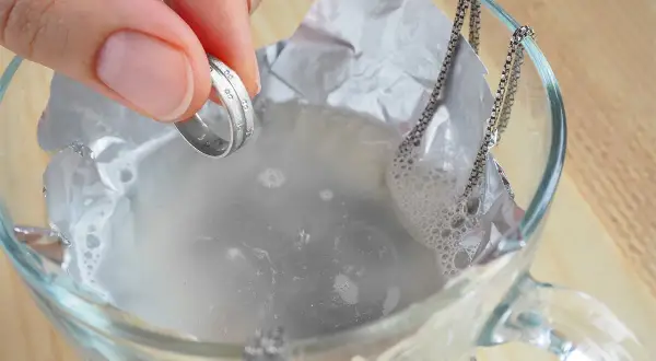Dłoń trzyma pierścionek nad szklanką z wodą i folią aluminiową, obok wisi łańcuszek.