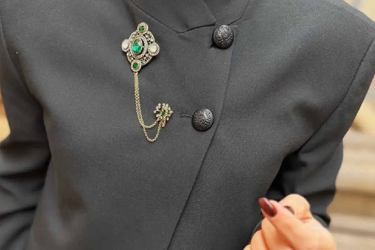 Elegancka broszka z zielonymi kamieniami i złotymi łańcuszkami dodaje szyku czarnej marynarce. Zobacz, jak nosić broszkę, by podkreślić stylizację.