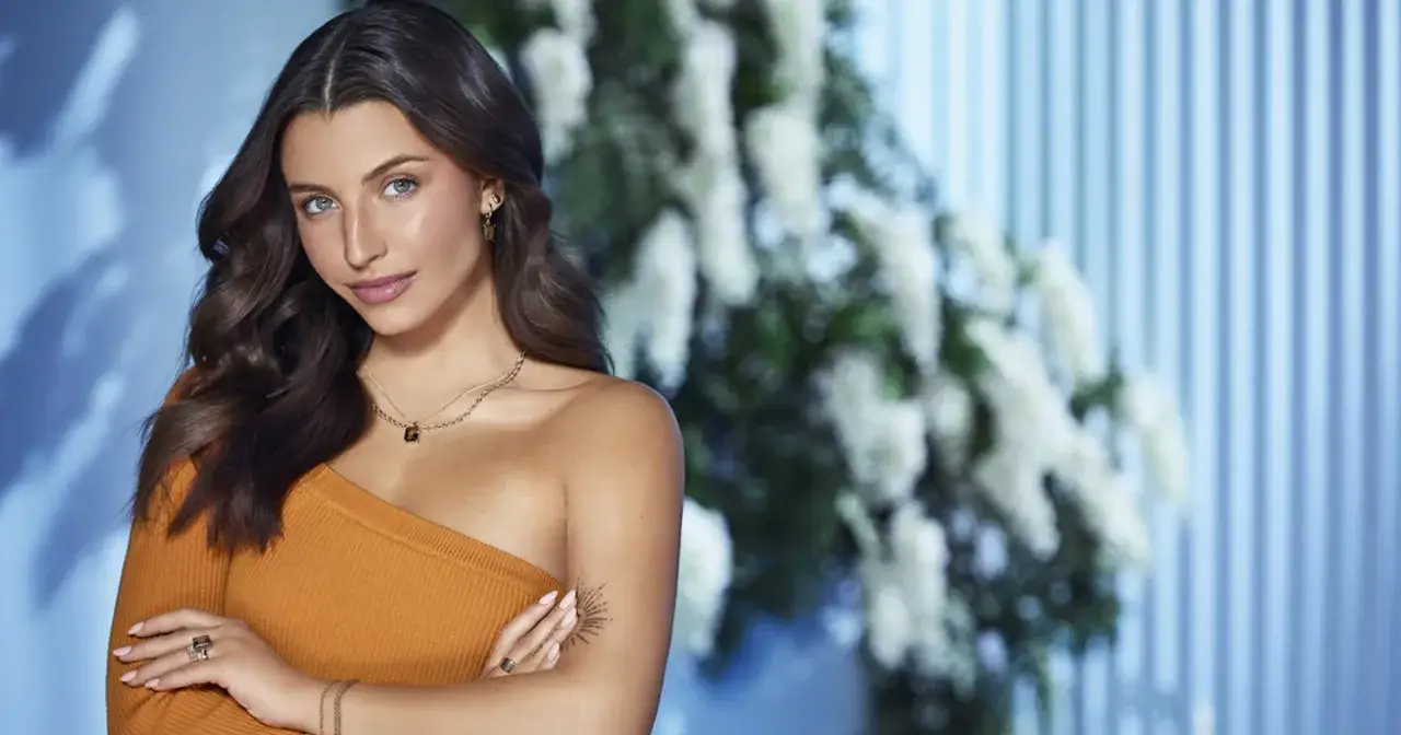 Młoda kobieta w pomarańczowej bluzce, kto reklamuje apart. Jej dłoń zdobią pierścionki i bransoletka.